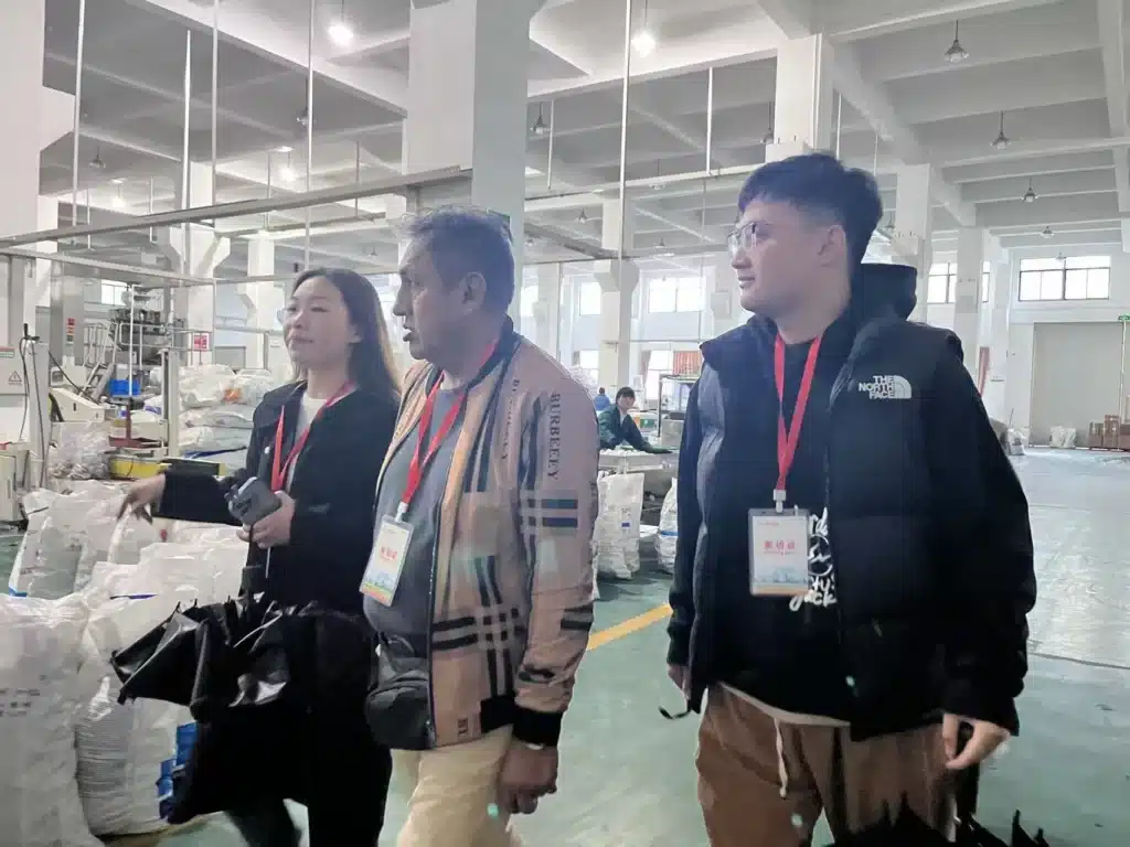 A group of people with identification badges are touring a factory with large bags and industrial equipment in the background.