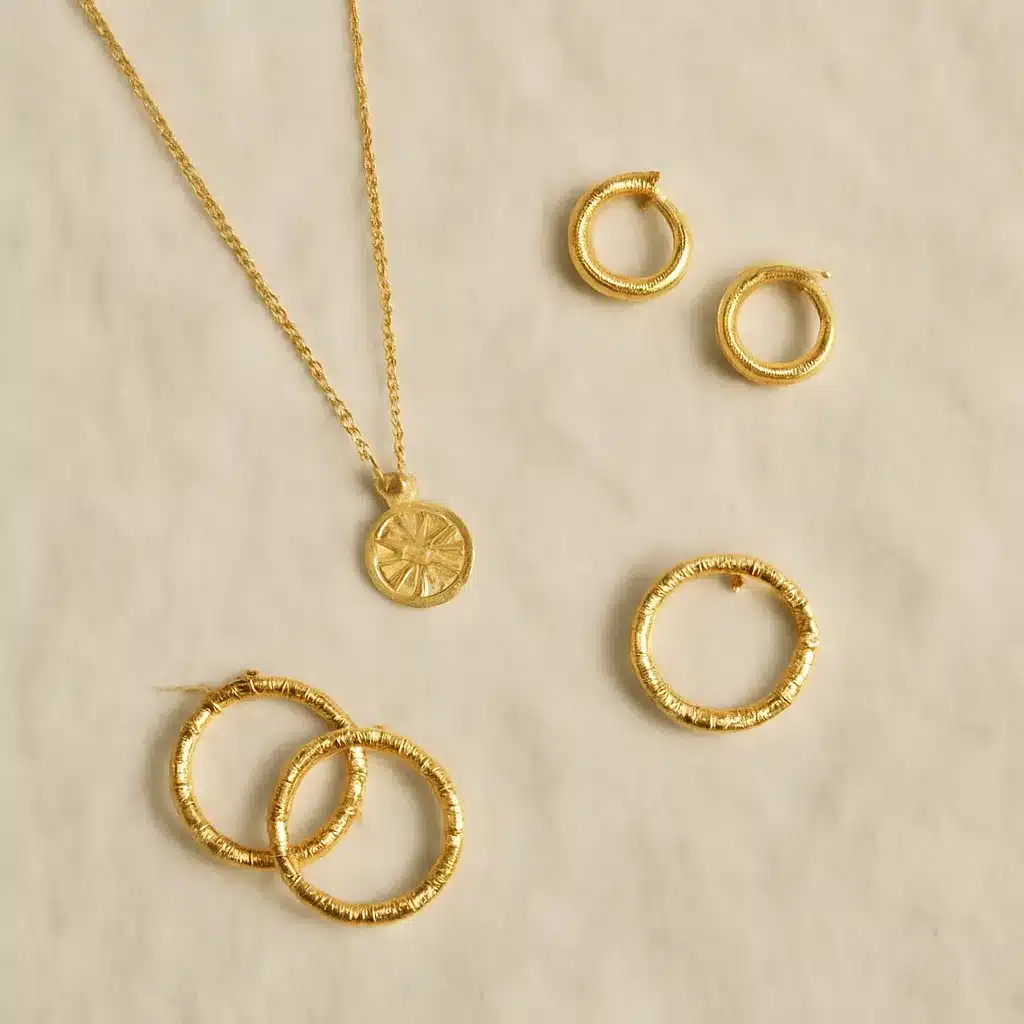 A gold jewelry set including a necklace with a circular pendant and three pairs of round hoop earrings displayed on a light background.