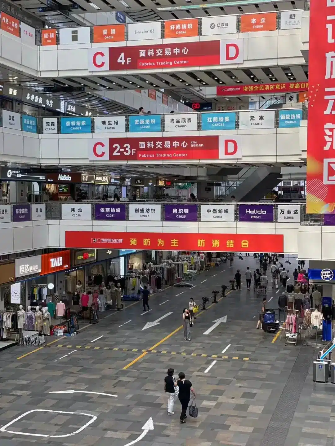 People walking through a multi-level fabrics trading center with numerous shops and signs, showcasing a bustling atmosphere.