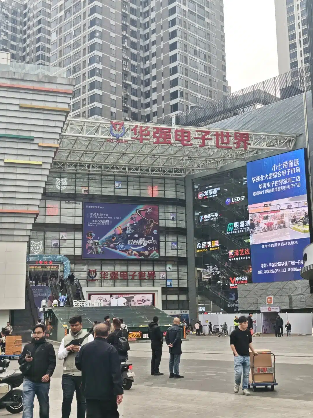 Large shopping complex exterior with electronic advertisements and signs, showing a busy urban area with people and high-rise buildings.