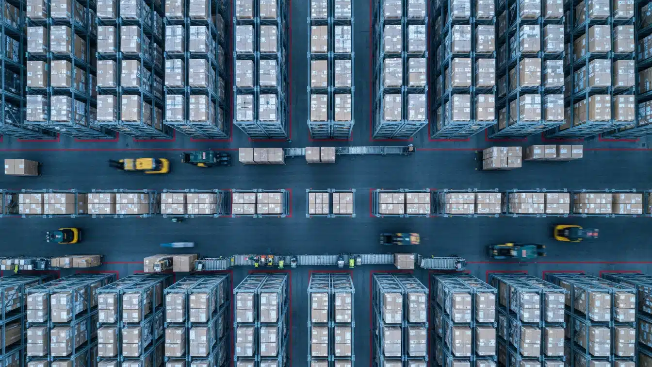 Aerial view of a large, organized warehouse with stacks of boxes and moving forklifts between aisles, showcasing efficient logistics.