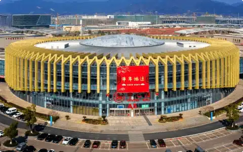 A modern circular building with yellow vertical slats and large glass windows, featuring a prominent red banner on its facade.