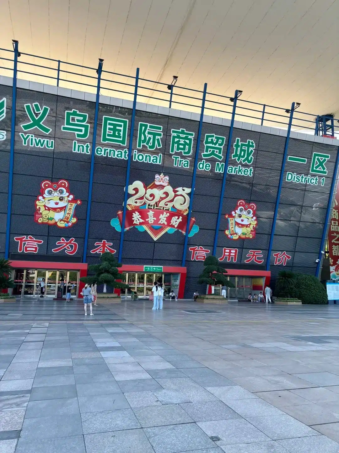 Entrance of Yiwu International Trade Market displaying the year 2023 with cartoon graphics, people walking in front, and signage in Chinese and English.