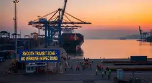 Cargo ship docked at a shipping port with cranes and containers during sunset, people in safety vests are visible on the dock.