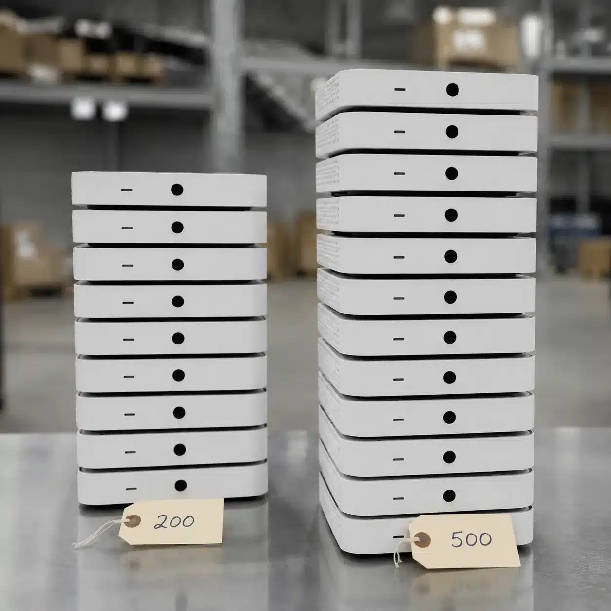 Hyper-realistic product photography, a close-up of two stacks of identical small consumer electronics on a warehouse table, left stack labeled with a subtle agent tag showing 200 units, right stack labeled with a factory tag showing 500 units, soft overhead lighting, clean industrial background, no text, no brand logo